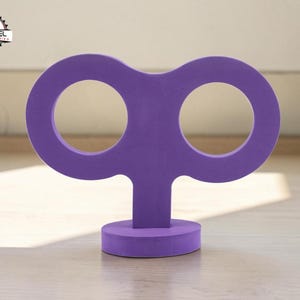 May include: A purple wind-up key-shaped decorative object. The object has two large circular openings and a small circular base. The object is made of a solid material and is sitting on a wooden surface.