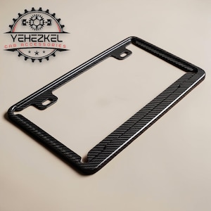 Könnte beinhalten: Ein schwarzer Kennzeichenrahmen aus Kohlefaser mit glänzendem Finish. Der Rahmen hat eine rechteckige Form mit abgerundeten Ecken und Befestigungslöchern. Das Logo "YEHEZKEL CAR ACCESSORIES" ist sichtbar.