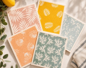 May include: Five square cellulose sponge cloths featuring various botanical patterns. The designs include white palm leaves on pink, yellow, and light blue backgrounds, white dandelion-like seeds on orange, and white coral-like branches on green.