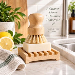 Op de afbeelding: Een lichtgekleurde houten afwasborstel met stijve haren staat op een rechthoekig wit stuk zeep, dat op een houten zeepbakje met latjes rust. De tekst A Cleaner Home A Healthier Tomorrow en PLASTIC FREE EVERYDAY is zichtbaar op de achtergrond.