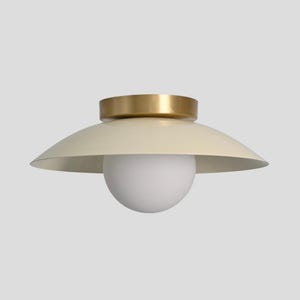 May include: A contemporary ceiling light. It features a cream-coloured, saucer-shaped shade and a gold-coloured top. A white, spherical light bulb hangs below the shade. Designed for indoor use.