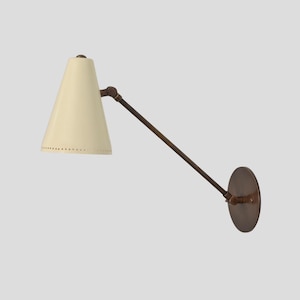 May include: Cream-colored cone-shaped wall sconce with a bronze-colored arm and circular base. The sconce has a series of small holes near the bottom edge of the shade. The arm extends from the base to the shade, allowing for adjustable positioning.