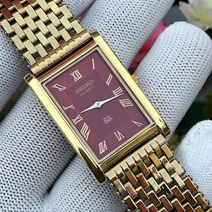 May include: A rectangular gold-tone Seiko Quartz watch with a deep burgundy dial featuring a subtle wavy pattern. The watch face displays gold Roman numeral hour markers, silver-tone hands, and a gold-tone metal link bracelet with a textured finish.