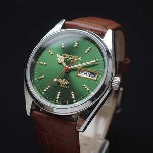 May include: A Citizen automatic wristwatch with a green dial, gold hands, and a day-date display. The watch has a silver-tone case and a brown leather strap. The dial features the text "CITIZEN AUTOMATIC" and "21 JEWELS".