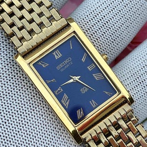 Może przedstawiać: Prostokątny zegarek na rękę Seiko Quartz z ciemnoniebieską tarczą, złotymi rzymskimi cyframi jako oznaczeniami godzin i pasującymi złotymi wskazówkami. Zegarek posiada polerowaną złotą kopertę oraz wieloogniwową metalową bransoletę w kolorze złota.