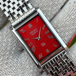 Könnte beinhalten: Eine rechteckige Seiko Quartz Armbanduhr mit einem leuchtend roten Zifferblatt mit strukturiertem Muster, silberfarbenen römischen Ziffern als Stundenmarkierungen und silbernen Zeigern. Die Uhr hat ein poliertes, mehrreihiges Gliederarmband.