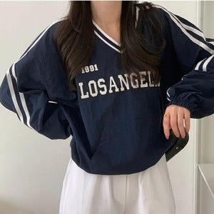 May include: Navy blue pullover with white stripes on the sleeves and a V-neck collar. The text "1991 LOSANGEL" is printed on the front in white. The sleeves are long and gathered at the wrists. The model is wearing white cargo shorts.