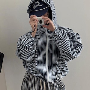 May include: A navy and white gingham hooded jacket with a front zipper, long sleeves, and a small rectangular white patch on the lower left side featuring black text. The jacket is paired with matching gingham pants and a navy baseball cap.