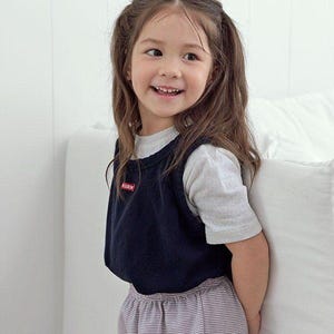 Puede incluir: Un niño lleva un chaleco azul marino con un pequeño logo rojo y blanco sobre una camiseta gris de manga corta. El niño también lleva pantalones a rayas. El texto "SN.STELLA" es visible en los pantalones.