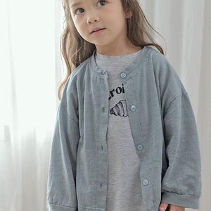 Op de afbeelding: Een kind draagt een lichtblauw vest met knopen over een grijze t-shirt. De t-shirt heeft een grafische print en de tekst "roi". De cardigan heeft bijpassende knopen. Onderaan de afbeelding staat de tekst "SN.STELLA".