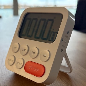 Könnte beinhalten: Ein weißer digitaler Küchentimer mit rechteckigem Design und integriertem Ständer. Der Timer zeigt "00:00" auf dem Bildschirm an und verfügt über Tasten für Stunden, Minuten und Sekunden. Die "ST/SP"-Taste ist rot. Der Timer steht auf einer Holzoberfläche.