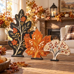 May include: Three decorative wooden leaf-shaped ornaments standing upright. The largest is black with orange flowers, the middle is orange with white berries, and the smallest is cream with red and orange leaves, all featuring painted botanical details.