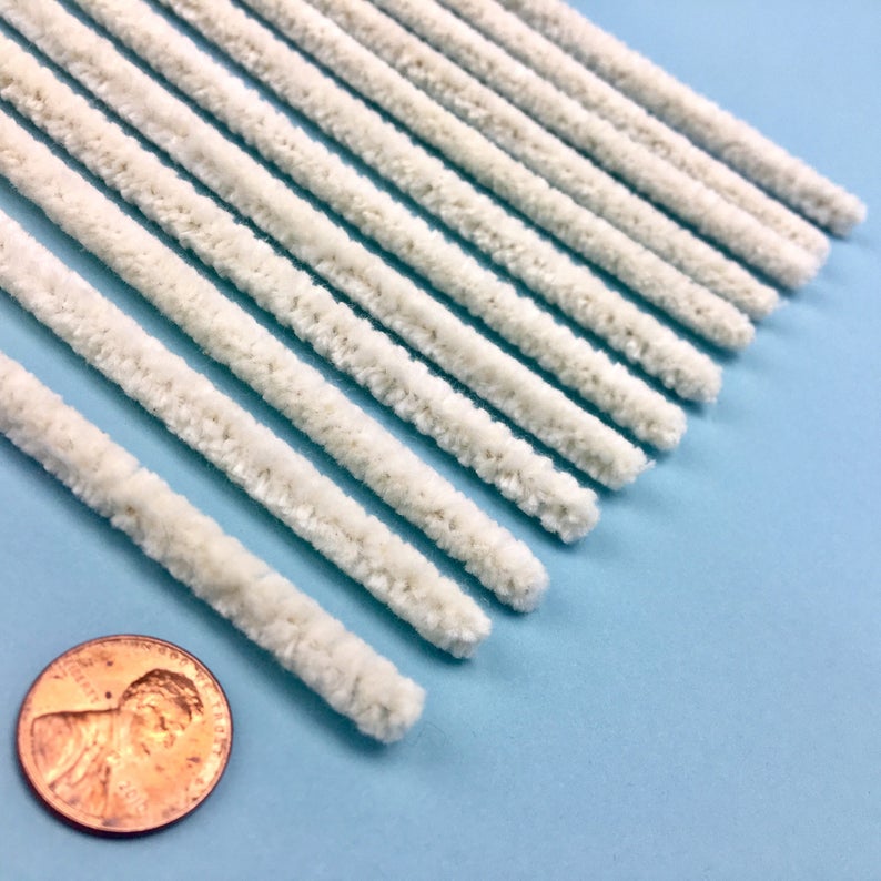 May include: A collection of off-white chenille stems, or pipe cleaners, displayed diagonally against a light blue backdrop. A penny is included for scale. These craft items are commonly used for various arts and crafts projects.