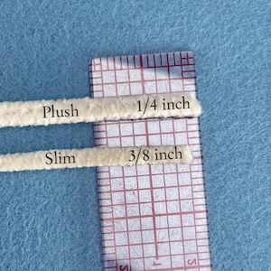 May include: Two beige pipe cleaners are shown against a light blue background, one labelled "Plush" and measuring 0.64 cm, the other "Slim" at 0.95 cm. A clear ruler with red grid lines is positioned next to the pipe cleaners.