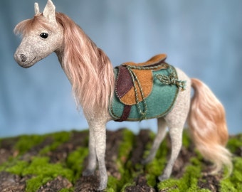 May include: A grey plush horse with a long, pink mane and tail. The horse is wearing a green and brown saddle with a brown leather strap.