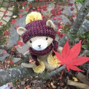 May include: A plush brown bear wearing a purple knitted hat with a yellow pom-pom and a matching scarf. The bear is sitting on a tree branch with a red maple leaf behind it.
