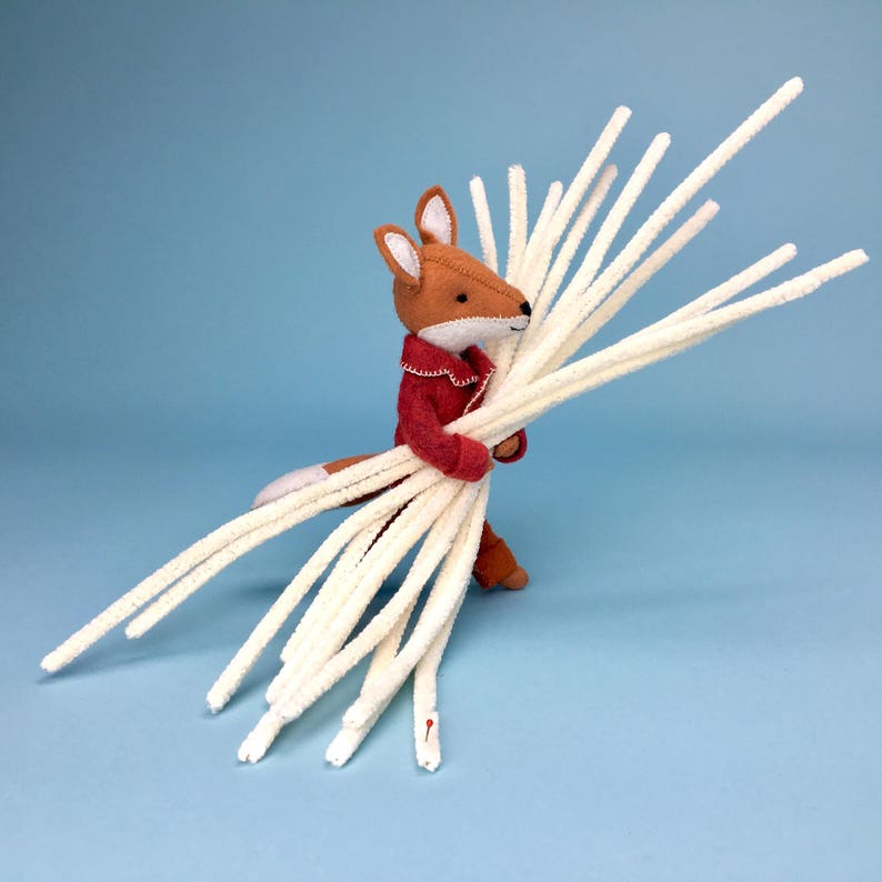 May include: A handcrafted felt fox toy in a red jacket, carrying a large bunch of cream-coloured pipe cleaners. The fox features brown and white fur, with a white chest and tail tip. The background is a solid light blue.