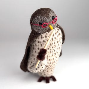 May include: A felted owl figurine featuring a dark brown head and wings with a cream-colored body covered in brown spots. The owl wears small red circular glasses, has a yellow beak, and holds a thin wooden pointer stick in one wing.