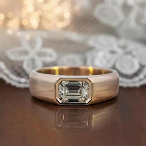 May include: A gold ring with a rectangular, emerald-cut diamond set in a bezel. The ring has a thick band and a polished finish. The background is blurred, with soft, warm lighting.