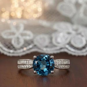 Könnte beinhalten: Ein silberfarbener Ring mit einem großen, runden, facettierten dunkelblauen Edelstein in der Mitte. Die Ringschiene ist auf beiden Seiten zweigeteilt, wobei jede Reihe mit kleinen, klaren, funkelnden Steinen entlang der sichtbaren Schiene besetzt ist.