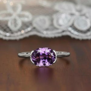 Può includere: Un anello d'argento con una grande pietra preziosa ovale viola. La pietra preziosa è tenuta in posizione da una montatura d'argento che si curva attorno alla pietra. Lo sfondo è sfocato.