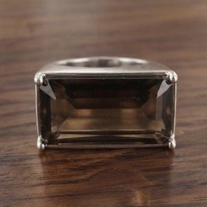 May include: A silver-toned ring featuring a large, rectangular, faceted smoky quartz gemstone set horizontally. The stone is held in place by four small prongs at each corner, and the band has a smooth, polished metallic finish.