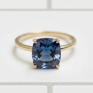 Pode incluir: Um anel de ouro com uma grande pedra preciosa azul escura de corte quadrado. A pedra é engastada com quatro garras, e a banda é de ouro polido. O anel é exibido sobre um fundo branco.