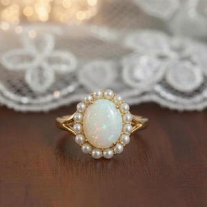 Könnte beinhalten: Ein goldener Ring mit einem ovalen Opal-Edelstein, umgeben von kleinen Perlen. Der Opal hat eine milchig-weiße Farbe mit schimmernden Reflexen. Der Ring hat ein filigranes Design mit geteiltem Schaft.