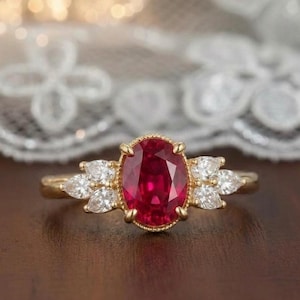 May include: A gold ring featuring a central oval-cut deep red ruby gemstone held in a prong setting, flanked on each side by three small pear-cut white diamonds. The band is crafted from polished yellow gold with a delicate beaded metal texture.