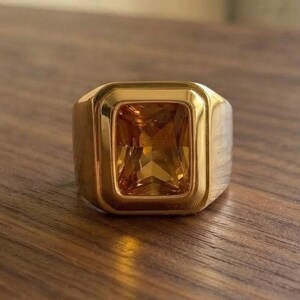 May include: A gold-toned ring featuring a large, rectangular, amber-colored gemstone set in a smooth, polished metal bezel. The ring has a thick, solid band that tapers slightly toward the back, showcasing a clean, reflective surface throughout.