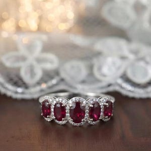 May include: A silver-toned ring featuring five oval-cut deep red rubies arranged in a horizontal row. Each ruby is surrounded by a halo of small, clear, round-cut stones set into the metal band, creating a continuous, sparkling, multi-stone design.