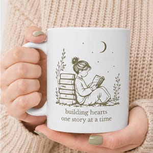 May include: White ceramic mug featuring a line drawing of a person reading a book, with a stack of books and a crescent moon. The mug has the text "building hearts one story at a time."