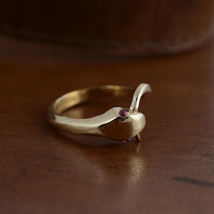 May include: A gold-colored ring in the shape of a snake, with a curved head and tail. The snake's head features two small, round, red gemstones. The ring is set against a dark brown wooden surface.