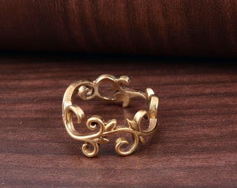 May include: A gold-colored ring with an intricate floral design. The ring features swirling vines and leaves, creating a delicate and ornate appearance. The ring is set against a dark brown wooden surface.