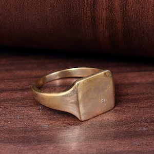 Pode incluir: Um anel de sinete dourado com uma face quadrada. O anel é feito de um metal com uma aparência ligeiramente envelhecida. O anel está sobre um fundo castanho escuro.
