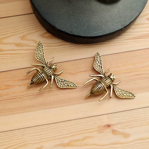 Könnte beinhalten: Zwei goldfarbene Bienenornamente mit detaillierten Flügeln und Körpern. Die Bienen, mit sichtbaren Beinen und Fühlern, liegen auf einer hellbraunen Holzoberfläche. Im Hintergrund befindet sich ein dunkles, rundes Objekt.