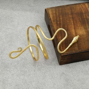 Peut inclure: Manchette de bras dorée en forme de serpent. Le bracelet s'enroule autour du bras avec un corps enroulé et une tête de serpent détaillée. Le bijou est présenté sur une surface en bois.