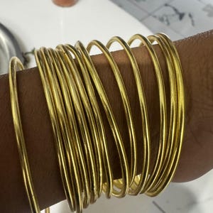 Pode incluir: Uma coleção de pulseiras douradas empilhadas em um braço. As pulseiras são finas, redondas e têm um acabamento brilhante. As joias são exibidas no braço de uma pessoa, mostrando a cor e o design das pulseiras. Um acessório simples e elegante.