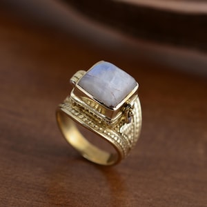 Pode incluir: Um anel dourado com uma pedra preciosa quadrada e clara. O anel tem uma banda ornamentada e texturizada e uma moldura quadrada em torno da pedra preciosa. A pedra preciosa tem uma cor azul clara e branca.