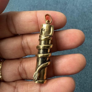 May include: A cylindrical, antique gold-colored pendant with a snake wrapped around it. The pendant has a small loop at the top and a detailed snake head at the bottom. The pendant is held in a hand.