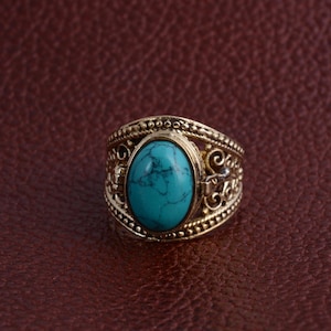 Pode incluir: Um anel dourado com uma pedra ovalada de cor turquesa. O anel tem um design ornamentado com filigranas e elementos decorativos em torno da pedra e da banda. O anel está sobre um fundo castanho.