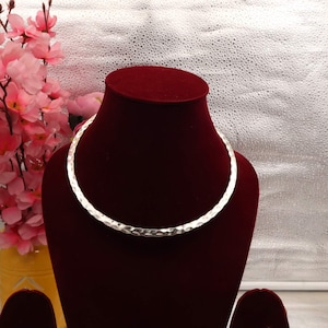 May include: A silver-colored hammered metal collar necklace displayed on a red velvet bust. The necklace has a textured surface and a simple, elegant design. Pink flowers and a yellow vase are in the background.