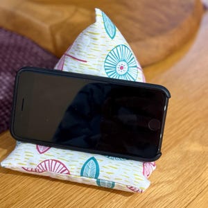 Puede incluir: Un smartphone negro descansa sobre un soporte de almohada estampada. La almohada presenta un fondo blanco con diseños de hojas y círculos en color turquesa, rosa y amarillo. El soporte está diseñado para sostener un teléfono para una visualización manos libres.