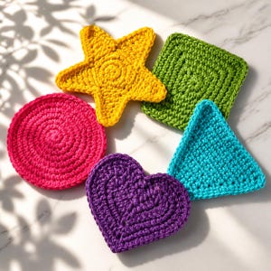 Puede incluir: Colección de posavasos de ganchillo hechos a mano y coloridos en varias formas: una estrella amarilla, un cuadrado verde, un triángulo azul, un corazón morado, un círculo rosa. Los posavasos están dispuestos sobre una superficie de mármol blanco.