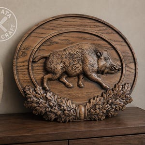 May include: An oval wooden wall plaque featuring a carved relief of a wild boar in the centre. The plaque is decorated with detailed oak leaves and acorns at the bottom. The wood has a natural brown grain finish and a circular logo in the corner.