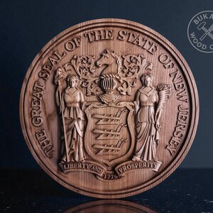 May include: A round, wooden carving of the Great Seal of the State of New Jersey. The seal features detailed relief carvings of figures, text, and the date 1776. The wood has a natural, warm brown tone. The text reads "Liberty and Prosperity".