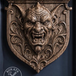 Puede incluir: Una placa de pared de madera marrón con una talla en relieve detallada de una cara de gárgola gruñendo con orejas puntiagudas y colmillos visibles, rodeada de volutas. En la esquina inferior izquierda aparece el logo BUKATA WOOD CRAFT.