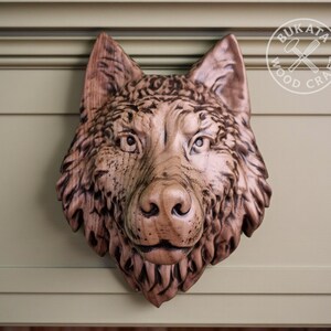 May include: A hand-carved wooden wolf head sculpture. The sculpture shows detailed features of the wolf's fur, eyes, and nose. The wood has a natural, warm brown tone. The sculpture is mounted on a light green surface.