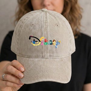 May include: A tan baseball cap with the embroidered word "Disney" in colorful letters. The hat has a curved brim and an adjustable strap at the back. The cap is made of a soft, worn material.