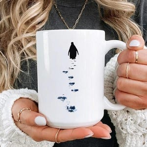 May include: White ceramic mug featuring a black penguin silhouette walking on a path of dark blue footprints. The mug has a large handle and a glossy finish, perfect for coffee or tea.